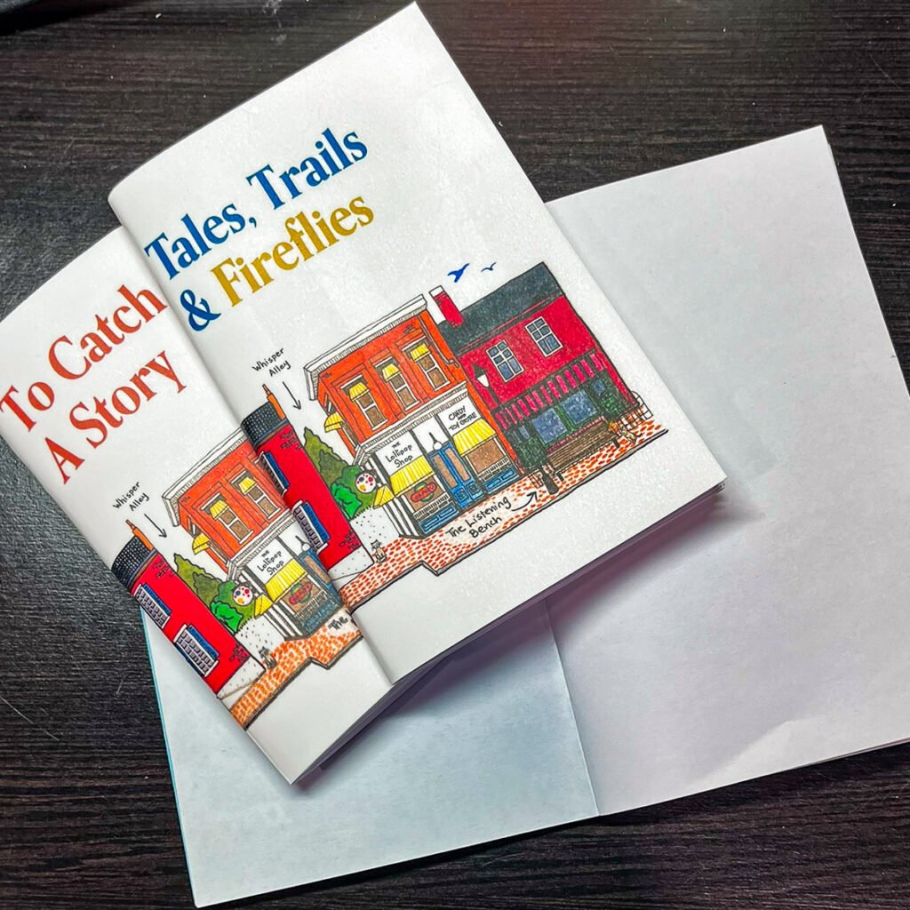 Two pocket-sized journals resting on a dark wood surface. The covers show the same illustrated downtown scene, but with different titles: “To Catch A Story” and “Tales, Trails & Fireflies.” A blank white envelope is placed beneath them.