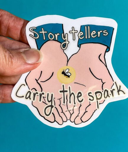Hand holding a large die-cut sticker that shows two open hands holding a glowing spark with a tiny firefly. The text reads “Storytellers Carry the spark.”