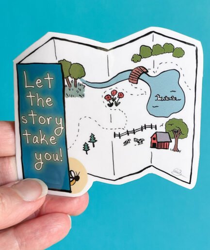 Hand holding a die-cut vinyl sticker shaped like an open map, with illustrated paths, trees, animals, and a pond. Text on the sticker reads: “Let the story take you!”