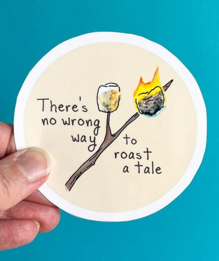 Hand holds a round sticker with two marshmallows on sticks, one burning, alongside the phrase “There’s no wrong way to roast a tale.”